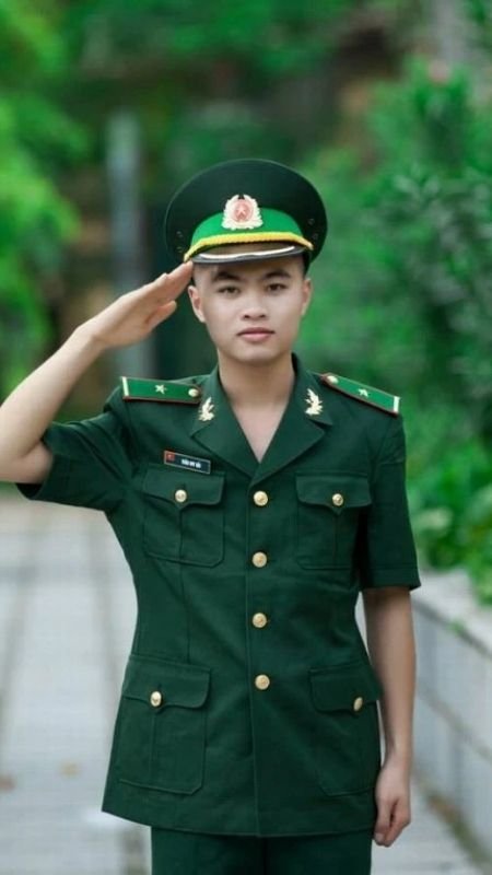 Người mặc quân phục màu xanh đang chào kiểu quân đội, đứng trên con đường có cây xanh xung quanh.