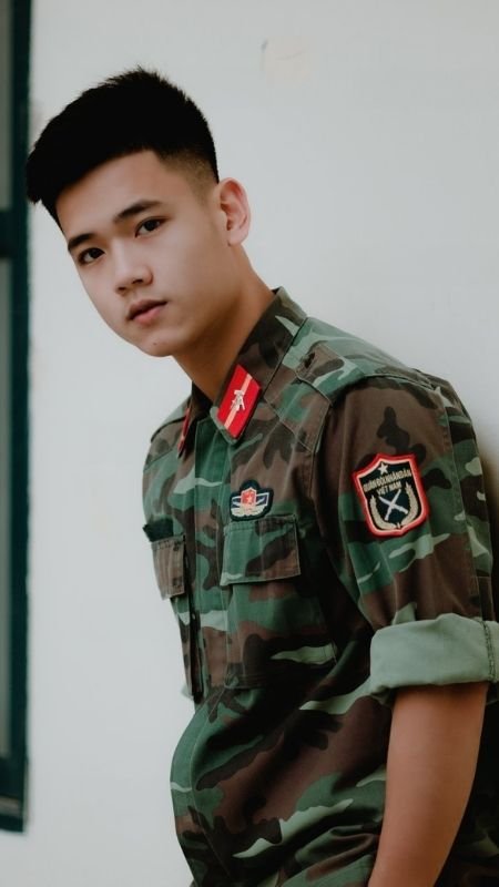 Người mặc quân phục đứng dựa vào tường, nhìn về phía trước, trên tay áo có gắn huy hiệu.