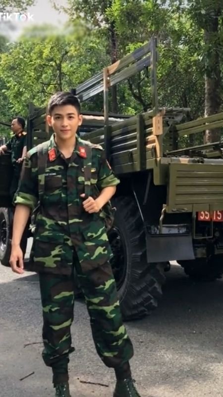 Người mặc quân phục đứng trước xe tải quân sự, khung cảnh rừng cây xung quanh.