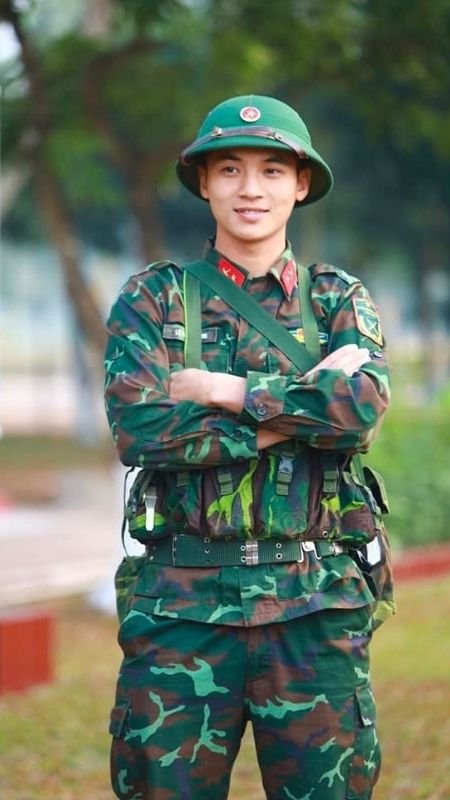 Người mặc quân phục đứng ngoài trời, cười, tay khoanh trước ngực, xung quanh là cây cỏ.