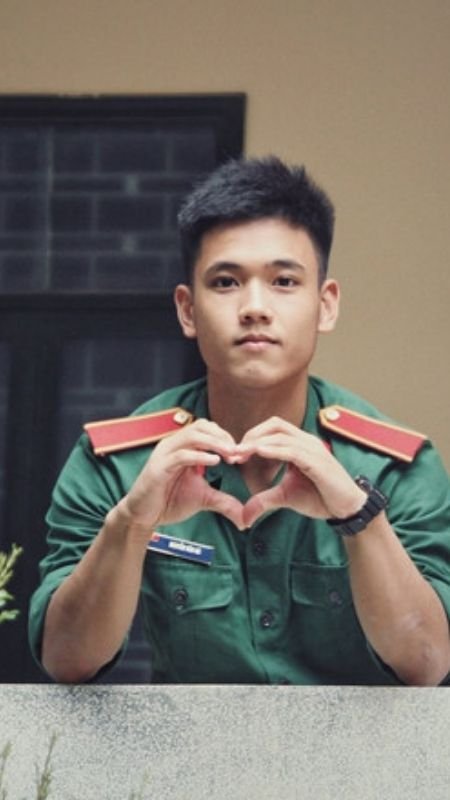 Người mặc quân phục màu xanh lá cây đang tạo hình trái tim bằng tay, đứng trước cửa sổ.