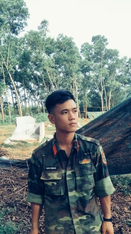 Một người mặc đồng phục quân đội đứng ngoài trời, nền là cây cối xanh tươi.