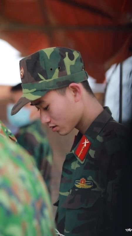 Người mặc quân phục rằn ri đứng nghiêng, đội mũ lưỡi trai, nhìn xuống, trong khung cảnh ngoài trời.