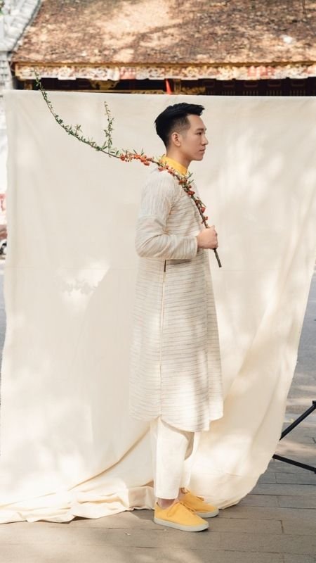 Người đàn ông mặc áo dài truyền thống, cầm cành hoa, đứng trước phông nền vải màu trắng ngà.