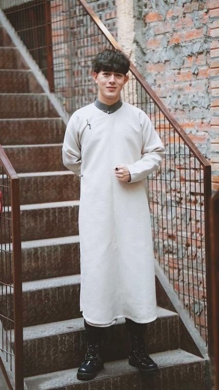 Người mặc áo dài trắng đứng trên cầu thang, tay để phía trước, nền là tường gạch thô.