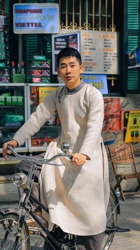 Người đàn ông mặc áo dài ngồi trên xe đạp trước cửa hàng, xung quanh có biển hiệu và sản phẩm.