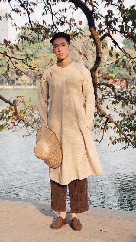 Người mặc áo dài truyền thống đứng cạnh hồ, tay cầm nón lá, xung quanh là cây cối và mặt nước.