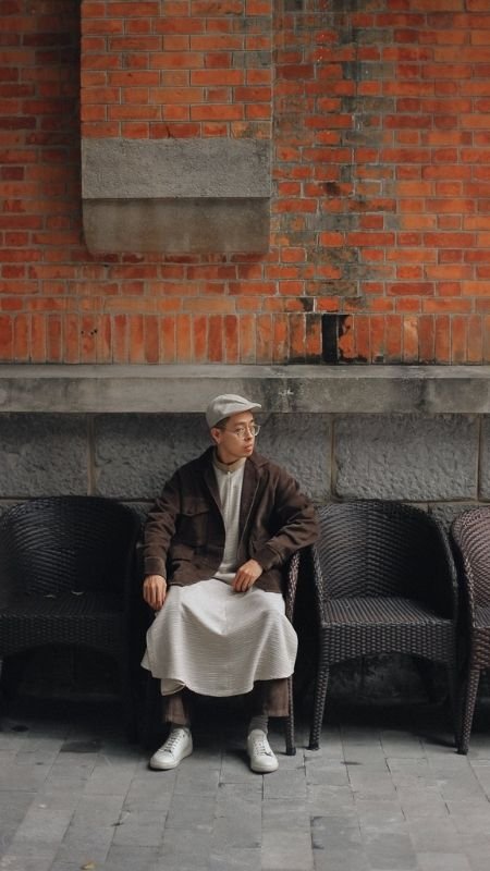 Người ngồi trên ghế mây, mặc áo khoác nâu và đội mũ xám, trước bức tường gạch đỏ.