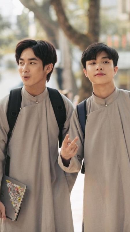 Hai người mặc áo dài truyền thống, đeo ba lô, đang đi bộ trên đường, một người cầm laptop.