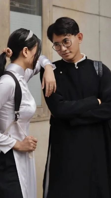 Hai người đứng trò chuyện, một người mặc áo dài trắng, người kia mặc áo dài đen, trước nền tường.