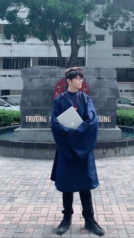 Người mặc trang phục truyền thống đứng trước tượng đài, tay cầm laptop, nền là cây xanh và tòa nhà.