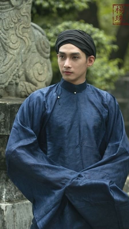 Người mặc trang phục truyền thống màu xanh đậm, đứng trước bức tượng đá cổ kính, xung quanh là cây xanh.