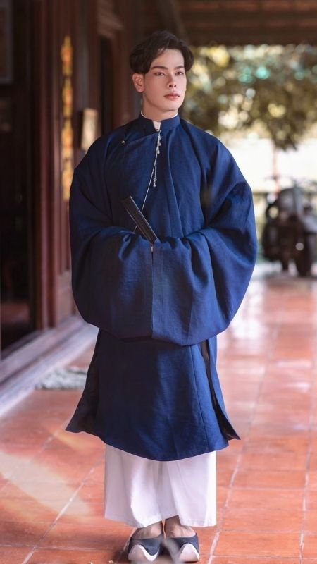 Người mặc áo dài xanh truyền thống, đứng trên hiên nhà lát gạch, tay cầm quạt, nền là cảnh vật tự nhiên.