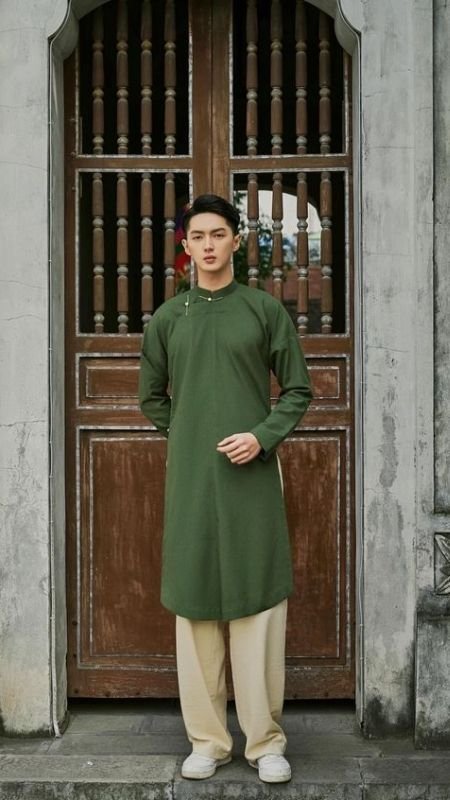 Người mặc áo dài xanh đứng trước cửa gỗ cổ điển, tay chắp phía trước, nhìn thẳng vào máy ảnh.