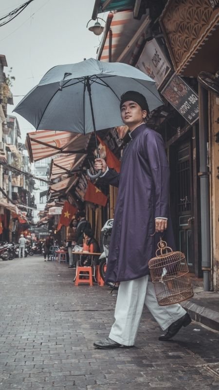 Người mặc áo dài, cầm ô và lồng chim, đi trên con phố nhỏ với nhiều cửa hàng và cờ đỏ sao vàng.