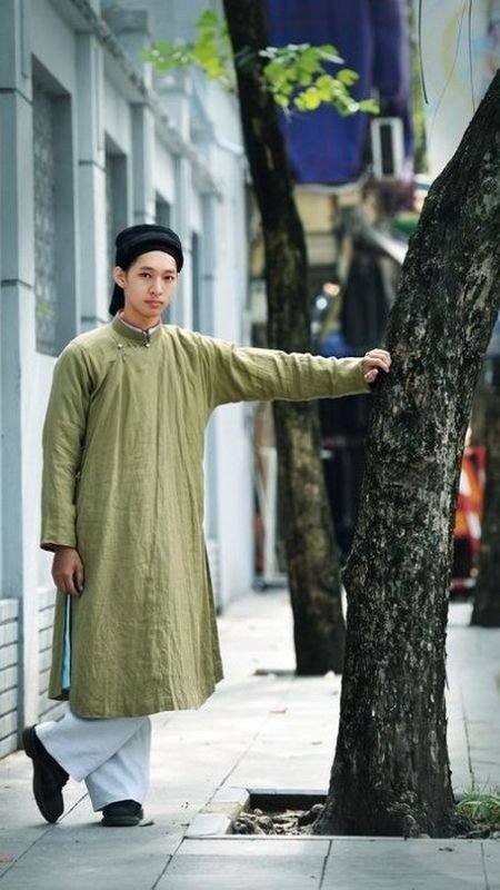 Người mặc áo dài truyền thống màu xanh lá nhạt, đội mũ đen, đứng cạnh cây trên vỉa hè phố.