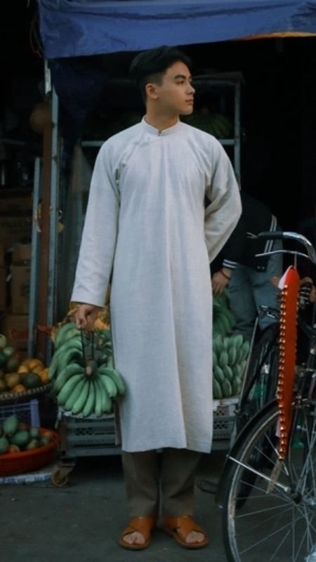 Người mặc áo dài trắng, cầm nải chuối xanh, đứng trước cửa hàng trái cây với xe đạp bên cạnh.