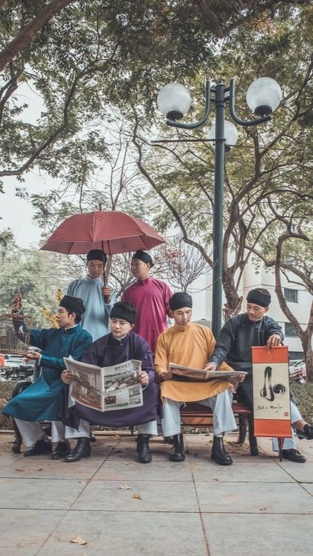 Nhóm người ngồi trên ghế công viên, mặc trang phục truyền thống, hai người cầm báo và người khác cầm ô.
