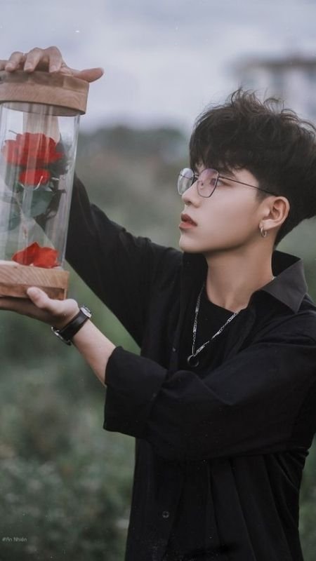 Người đeo kính cầm hộp gỗ chứa ba hoa hồng đỏ, nền cảnh đồng cỏ xanh mờ.