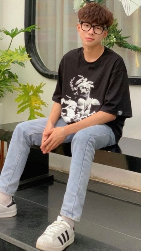 Người ngồi trên ghế, mặc áo thun đen in hình, quần jeans xanh nhạt, giày thể thao trắng, kính đen. Có cây xanh xung quanh.