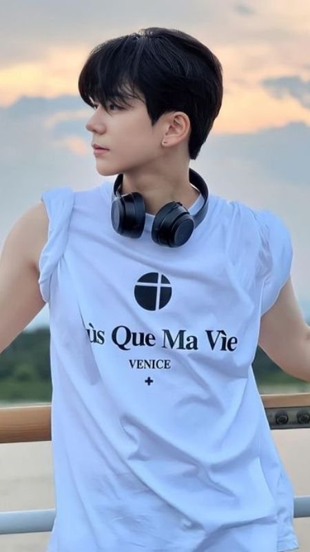 Người mặc áo phông trắng và đeo tai nghe, đứng ngoài trời với bầu trời hoàng hôn phía sau.