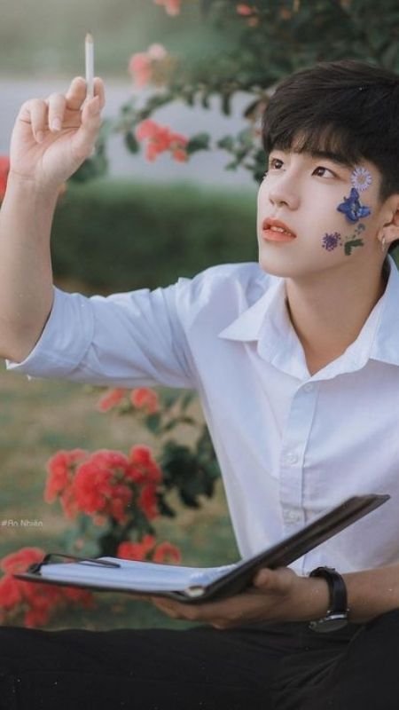 Người cầm bút và sổ, có hình hoa vẽ trên mặt, ngồi trước bụi hoa đỏ, ánh mắt nhìn lên trên.