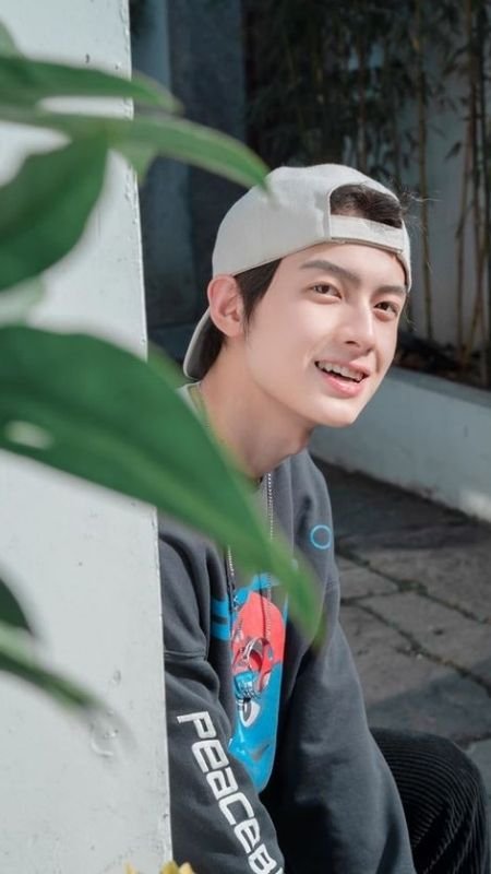 Người đội mũ lưỡi trai ngược, mặc áo dài tay, ngồi cạnh bức tường trắng, phía trước có lá cây xanh.