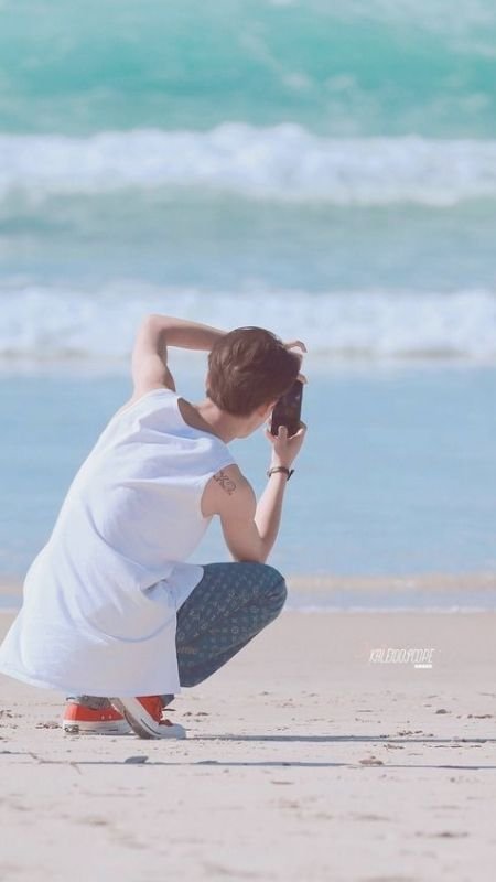 Người mặc áo trắng đang chụp ảnh trên bãi biển, với sóng biển phía sau.