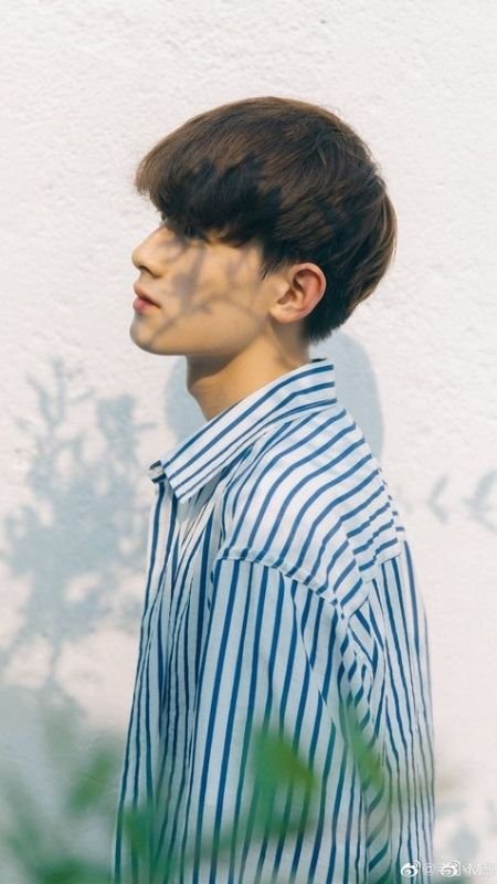 Người mặc áo sơ mi kẻ sọc xanh trắng, đứng nghiêng trước bức tường sáng màu, ánh sáng tạo bóng mờ trên mặt.