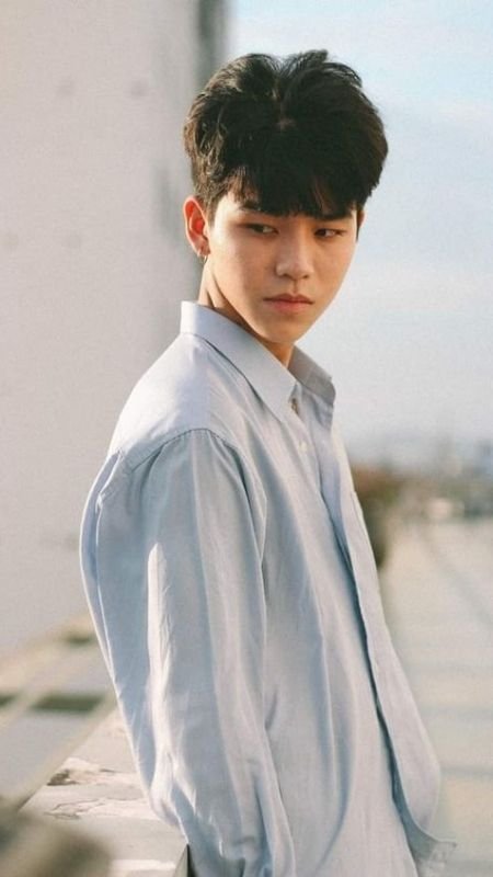 Người đàn ông mặc áo sơ mi đứng ngoài trời, nhìn sang bên, nền là bầu trời và tòa nhà.