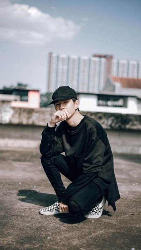 Người đội mũ đen ngồi trên sân thượng, mặc áo đen và giày caro, phía sau là tòa nhà cao tầng.