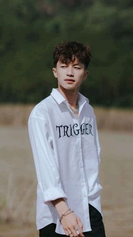 Người đàn ông mặc áo sơ mi trắng in chữ, đứng ngoài trời với phong cảnh thiên nhiên phía sau.