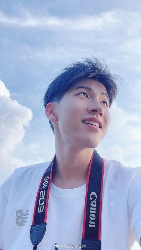 Người đang mỉm cười, đeo máy ảnh quanh cổ, đứng dưới bầu trời xanh với mây trắng.