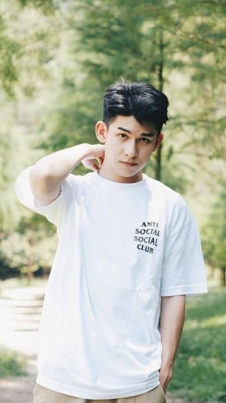 Người mặc áo phông trắng in chữ đứng trong khung cảnh cây xanh, ánh sáng tự nhiên chiếu sáng khuôn mặt.