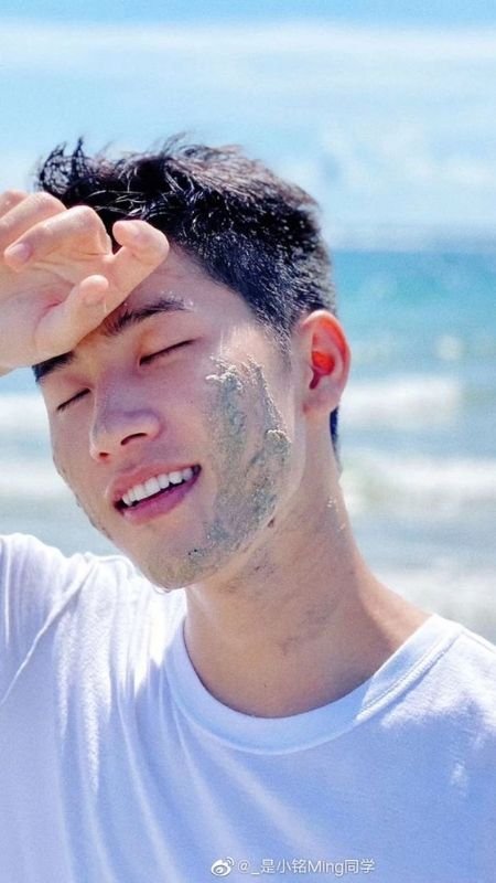 Người đàn ông mặc áo trắng đứng ngoài bãi biển, mỉm cười với cát dính trên mặt dưới ánh nắng.