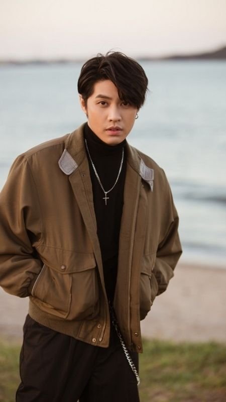 Người đàn ông mặc áo khoác nâu và áo cổ lọ đen, đứng trước bãi biển với bối cảnh hoàng hôn.