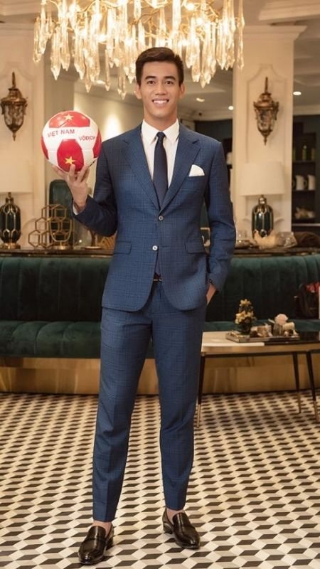 Người đàn ông trong bộ vest xanh cầm quả bóng đá Việt Nam, đứng trên nền gạch hoa, dưới đèn chùm sang trọng.