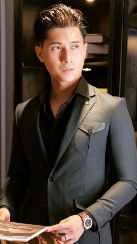 Người đàn ông mặc vest lịch lãm, cầm một quyển sách và nhìn xa xăm trong không gian nội thất sang trọng.