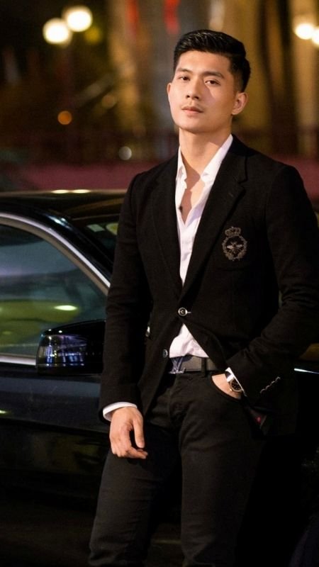 Người đàn ông mặc vest đen và áo sơ mi trắng đứng tựa vào xe hơi trong khung cảnh thành phố về đêm.