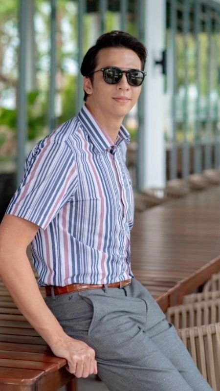 Người đàn ông đeo kính râm, mặc áo sơ mi sọc, ngồi trên ghế gỗ ngoài trời, xung quanh có cây xanh.