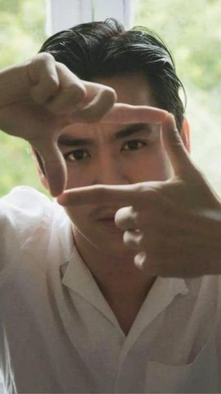 Người đàn ông mặc áo trắng tạo khung hình bằng ngón tay trước gương mặt, nền là cảnh thiên nhiên mờ ảo.