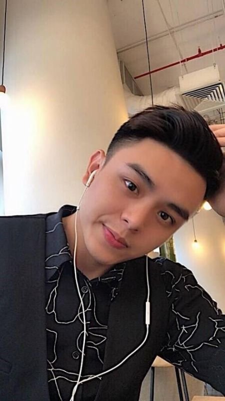 Người đàn ông trẻ đeo tai nghe, mặc áo sơ mi tối màu, ngồi trong quán cà phê, tay đặt lên đầu, nhìn vào máy ảnh.