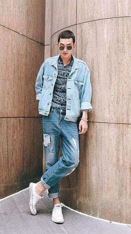 Người mặc áo khoác và quần jeans đứng dựa vào tường, mang kính râm và giày thể thao trắng.