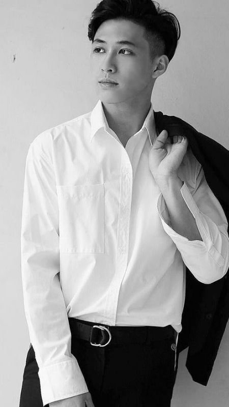 Người đàn ông mặc áo sơ mi trắng, khoác áo vest trên vai, đứng dựa vào tường, nhìn ra xa.