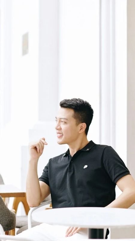 Người đàn ông mặc áo đen ngồi trên ghế, cười và nhìn sang trái, trong không gian sáng sủa với bàn ghế xung quanh.