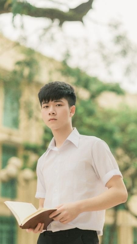 Người trẻ mặc áo sơ mi trắng đang cầm cuốn sách, nền là khung cảnh ngoài trời với cây xanh và tòa nhà.