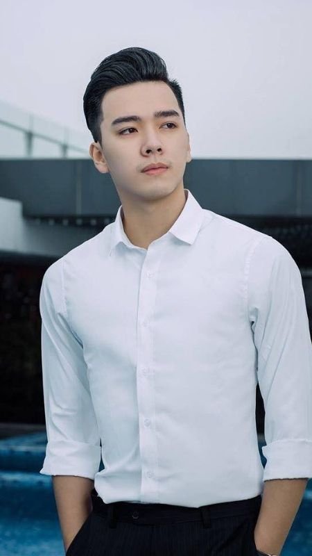 Người đàn ông trong áo sơ mi trắng đứng ngoài trời, phía sau là hồ bơi và kiến trúc hiện đại.