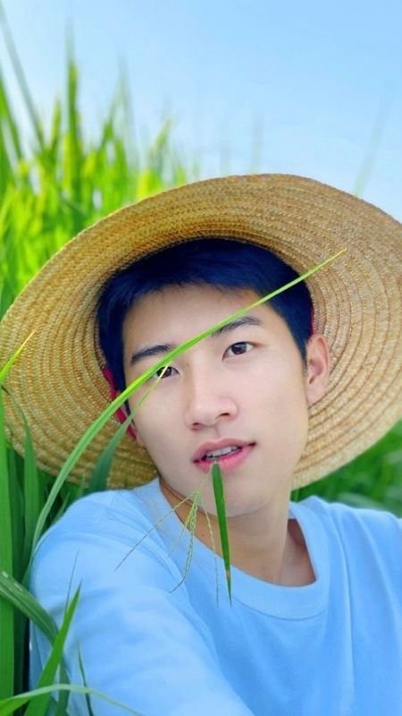 Người đội nón rộng vành, mặc áo xanh, đứng giữa cánh đồng cỏ xanh tươi dưới bầu trời trong xanh.