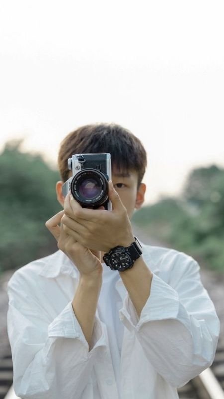 Người mặc áo trắng chụp ảnh bằng máy ảnh, đứng trên đường ray với phong cảnh thiên nhiên phía sau.
