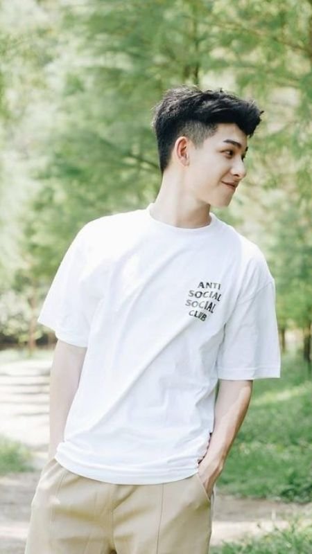 Người mặc áo thun trắng đứng ngoài trời, tay cho vào túi quần, nền là cây xanh mát.